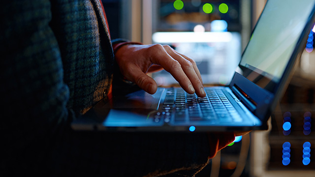A person in a dark sweater types on a laptop in a dimly lit server room.