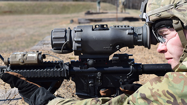 Soldier in camouflage, aiming through a high-tech scope mounted on a rifle, focused expression, outdoors on a training ground.