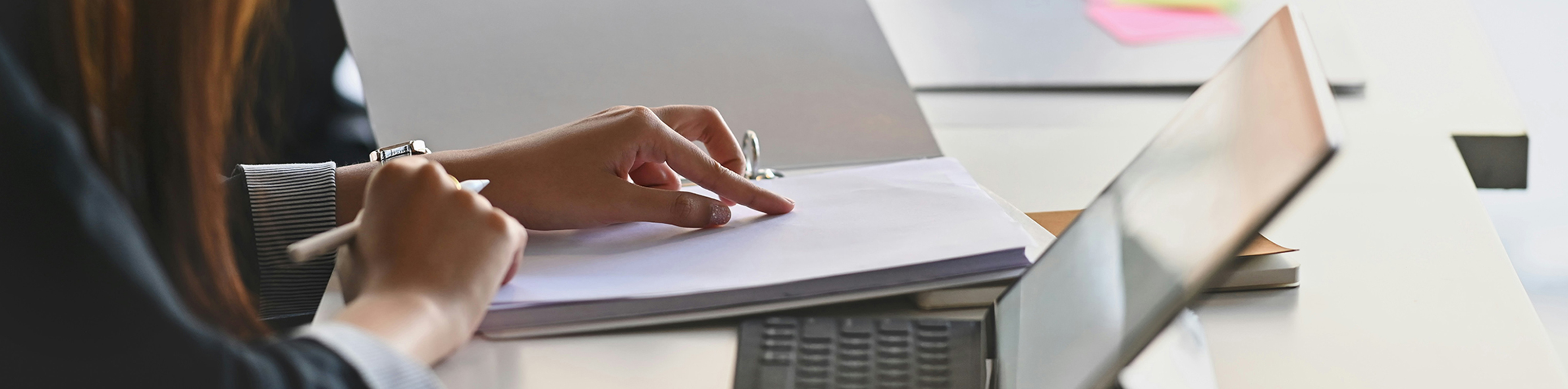 A woman sits at a desk, writing on a piece of paper with a focused expression.