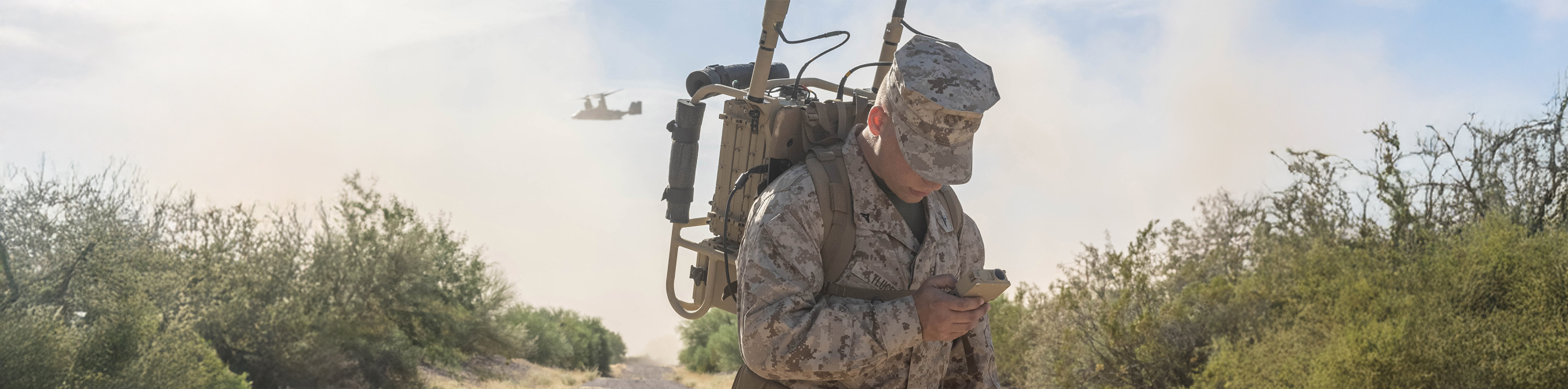 A soldier stands on a dirt road with a helicopter hovering in the background, showcasing a military environment.