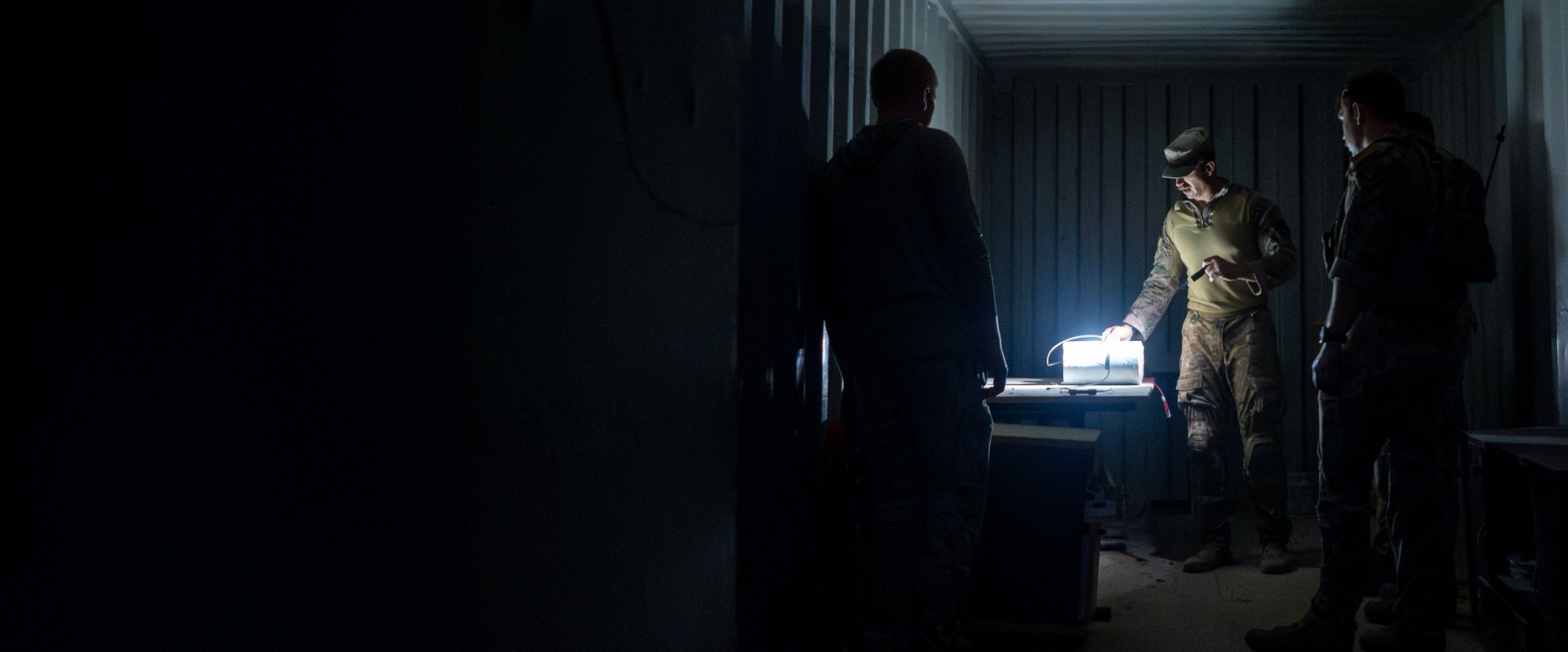 Two soldiers stand in a dimly lit room, illuminated by a single light source above them.