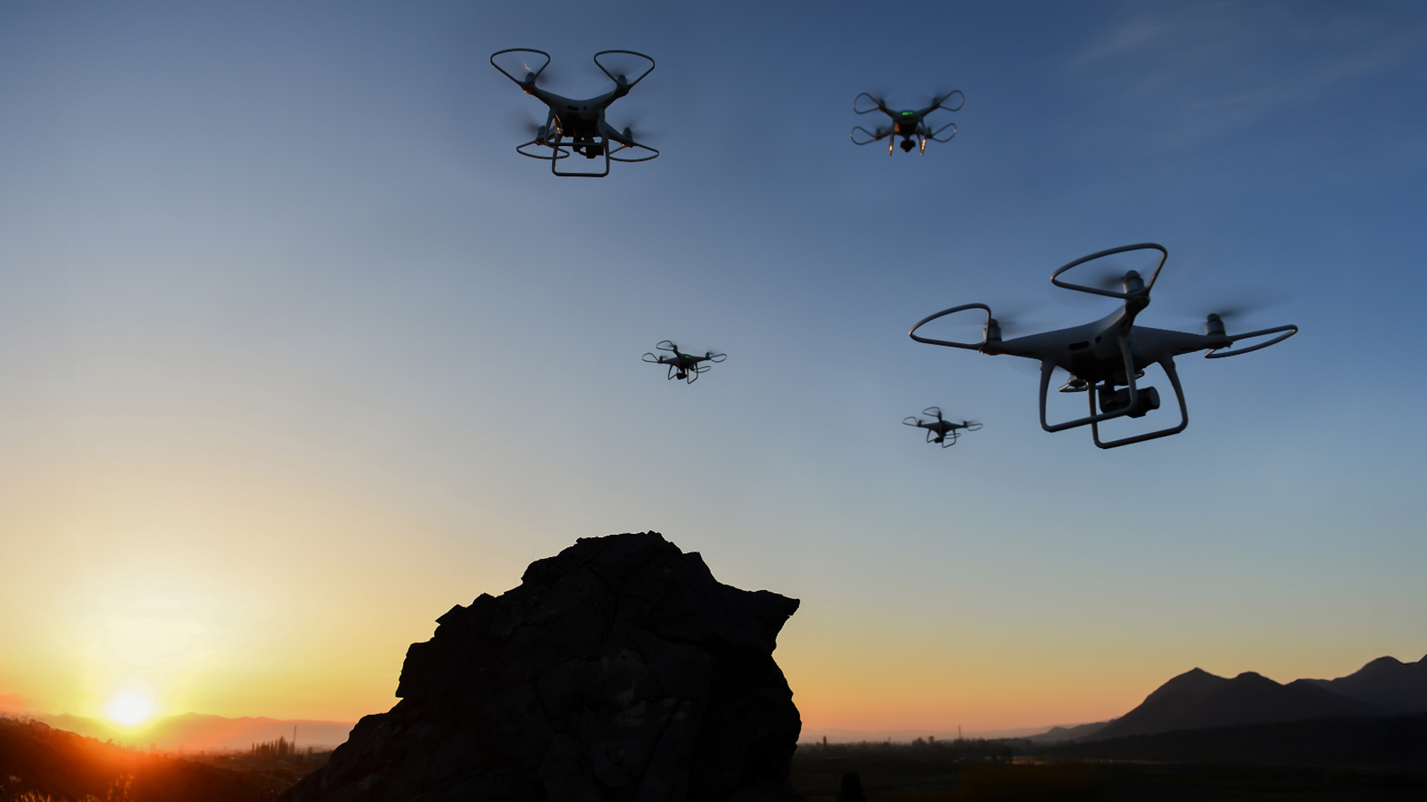 Four drones hover in the evening sky over a silhouetted landscape with a rocky foreground and distant mountains, set against a vibrant sunset.
