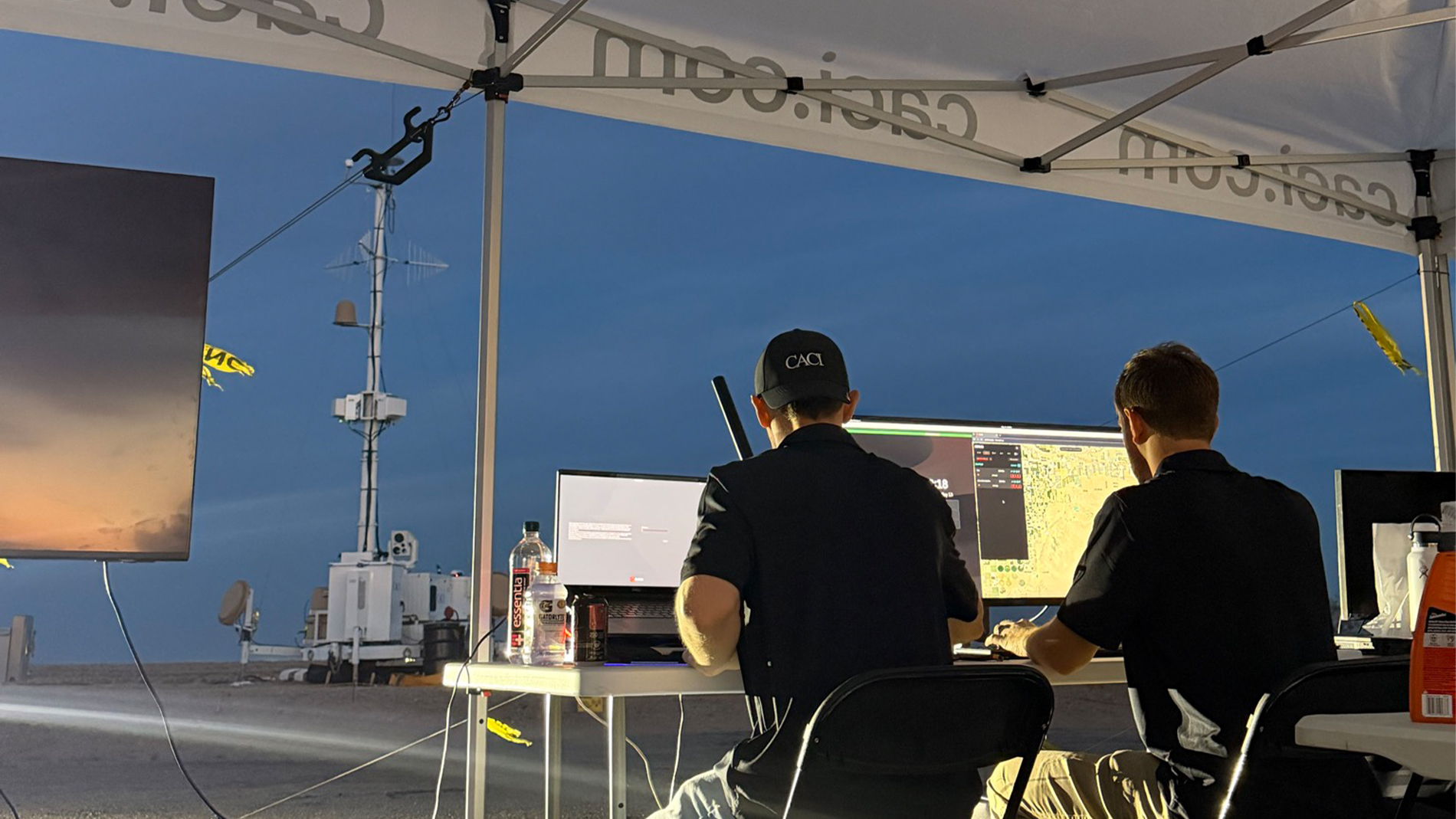 Two men seated at a table under a tent, working on laptops with focused expressions.
