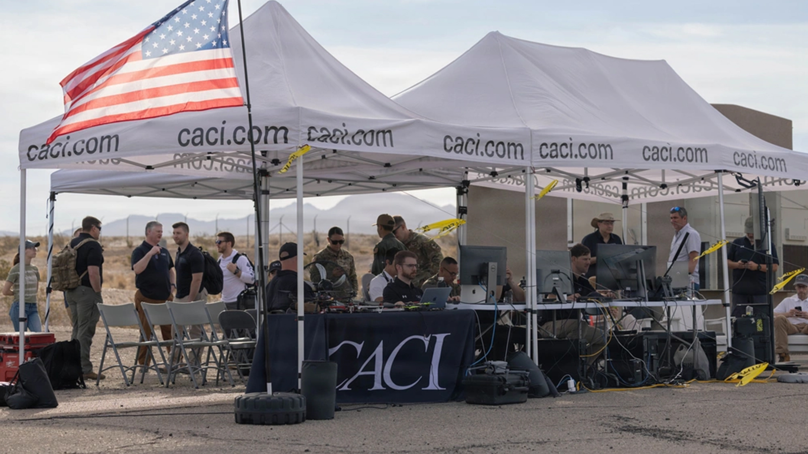 A group of people seated under a tent, with an American flag prominently displayed in the background.