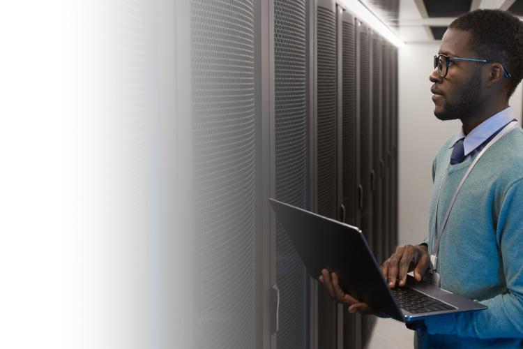 A man in a blue sweater stands in a server room, holding a laptop while surrounded by rows of servers.
