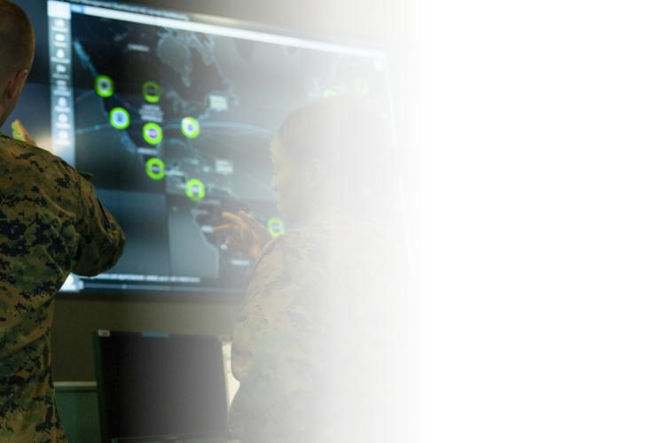 Two men in military uniforms observe a computer screen, engaged in a focused discussion or analysis.