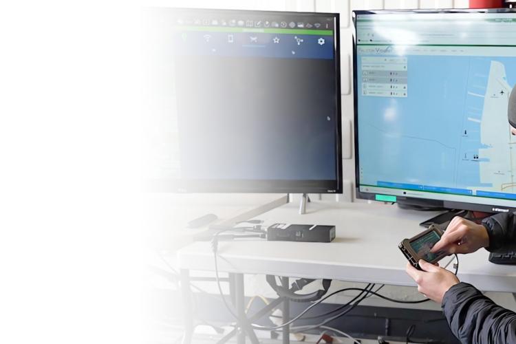 A man in a hat and black jacket sits at a desk, focused on two computer monitors in front of him.
