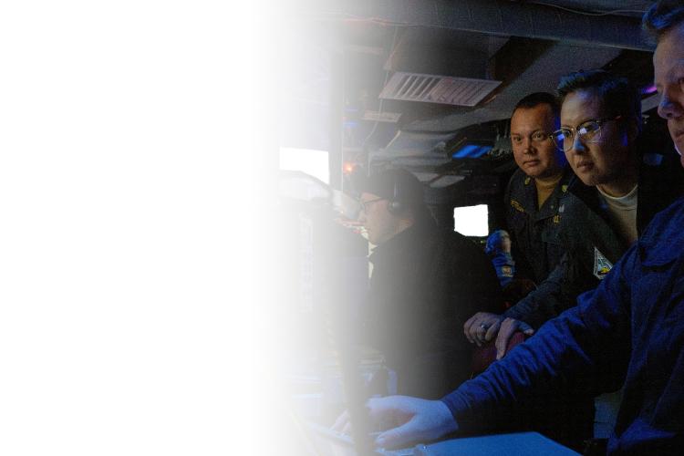 A group of men gathered in a room, focused on a laptop placed on a table in front of them.