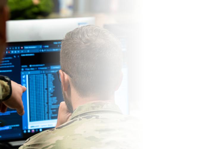 Two soldiers observe a computer screen, engaged in discussion or analysis of the displayed information.