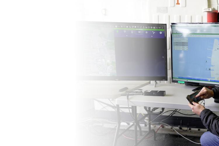 A man in a hat and black jacket sits at a desk, focused on two computer monitors in front of him.