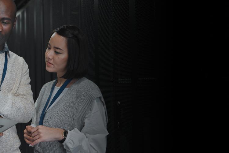 Two individuals examining a laptop in a server room filled with servers and networking equipment.