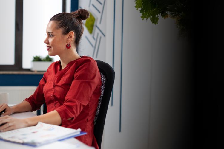 A woman sits at a desk, focused on her computer, with papers and a coffee cup nearby.