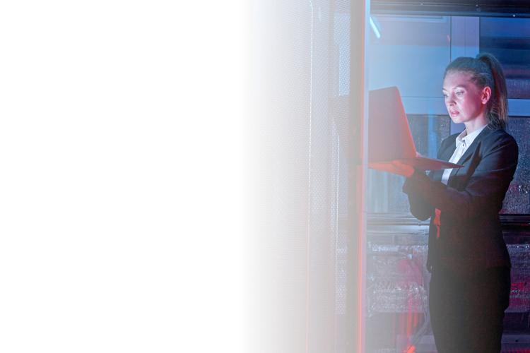 A woman stands in a server room, focused on a computer surrounded by rows of servers and blinking lights.