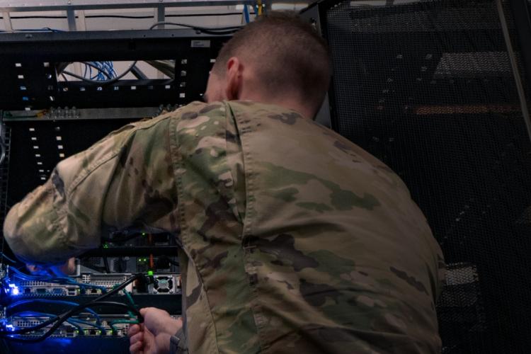 A soldier is configuring a server inside a data center, surrounded by rows of computer equipment and blinking lights.