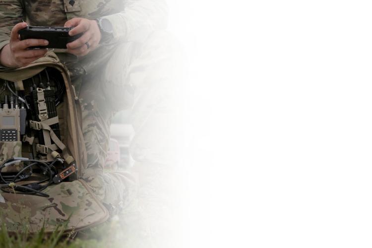 A man in camouflage gear stands holding a cell phone, focused on the device in his hand.