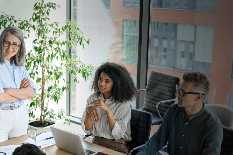 A woman stands at the front of a meeting room, addressing a group of seated people engaged in discussion.