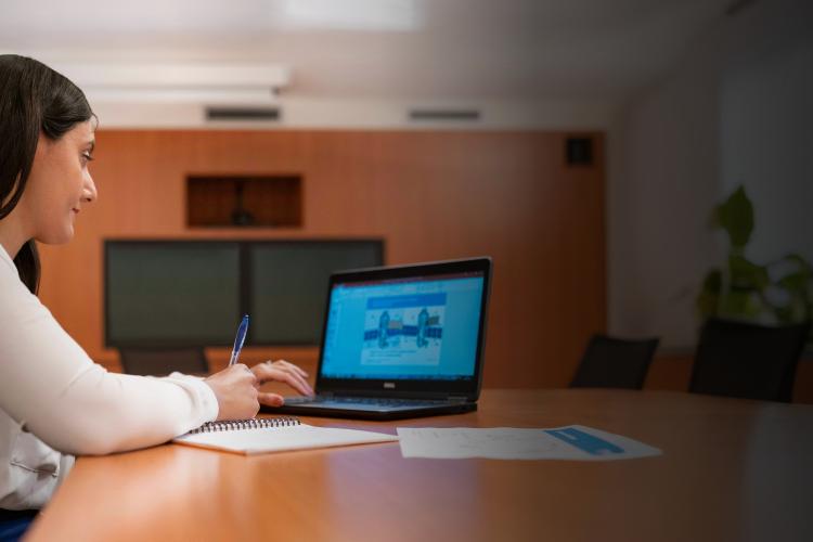 A woman seated at a table, focused on her laptop, with a calm expression in a well-lit room.
