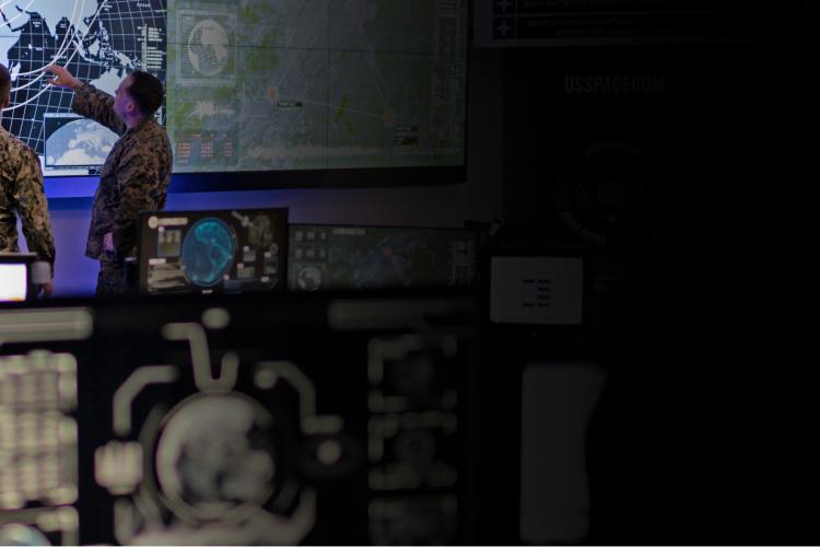 Two men in military uniforms stand in front of a large screen displaying information or visuals.