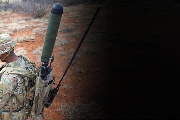 A soldier sits on the ground, holding a rifle, with a focused expression in a military setting.
