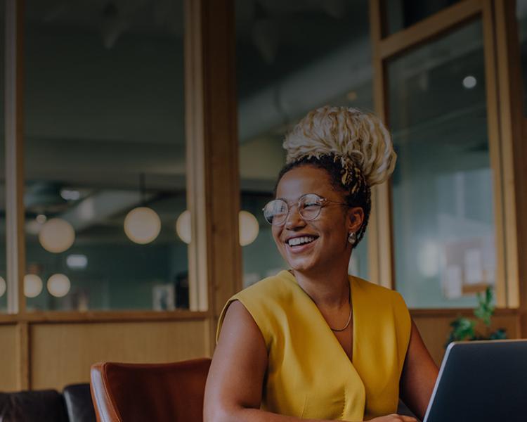 A woman seated at a table, focused on her laptop, with a calm expression in a well-lit room.