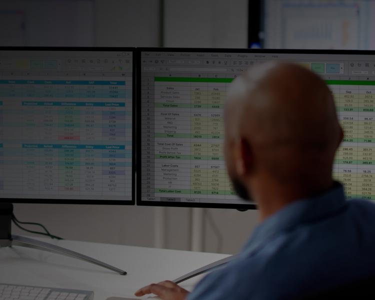 Two individuals seated at a desk, working together with multiple computer monitors in front of them.