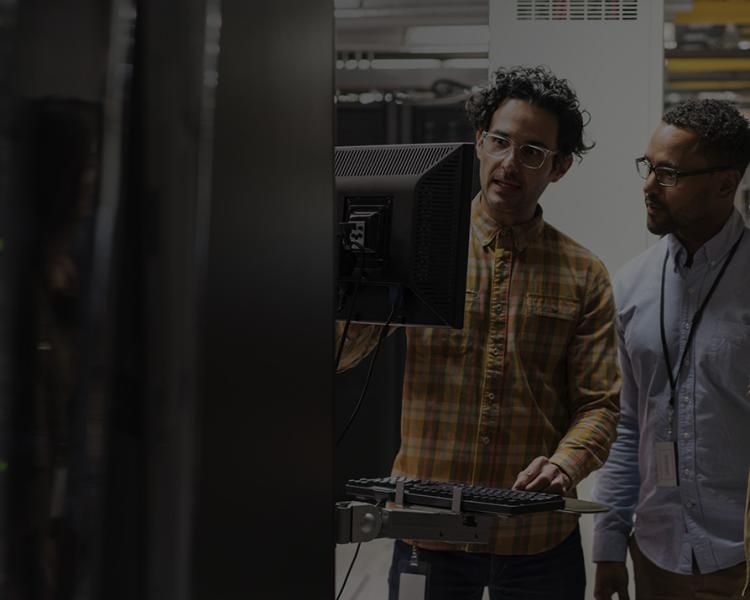 Three individuals examining a computer screen in a server room filled with racks of servers and blinking lights.