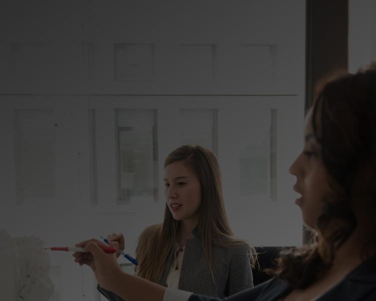 A woman writes on a whiteboard while another woman observes attentively.