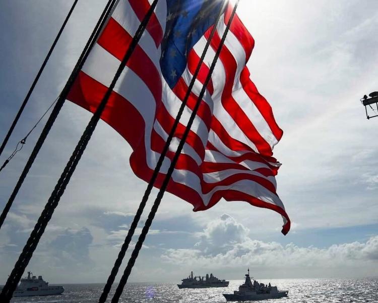 The deck of a ship displays the U.S. flag, billowing in the wind under bright sunlight.