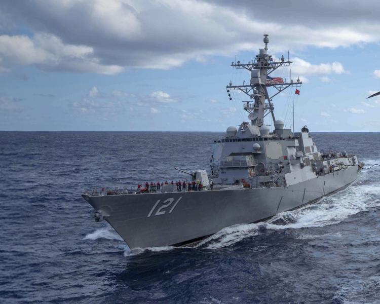 A navy destroyer sails through the ocean, viewed from an aircraft carrier's deck. The scene is under a partly cloudy sky.
