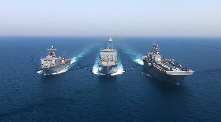 A formation of three military ships, including an amphibious assault ship, a destroyer, and a logistics support ship, steams across a calm ocean.