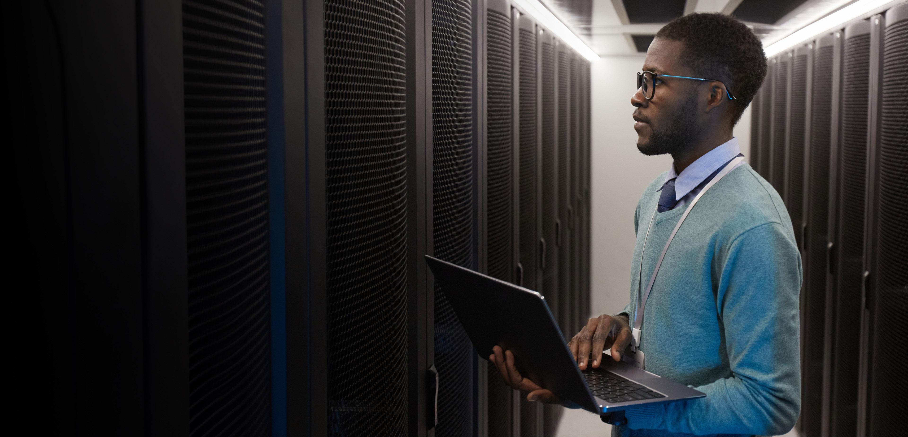 A man with glasses and a sweater stands in a server room, overseeing the array of servers and technology around him.