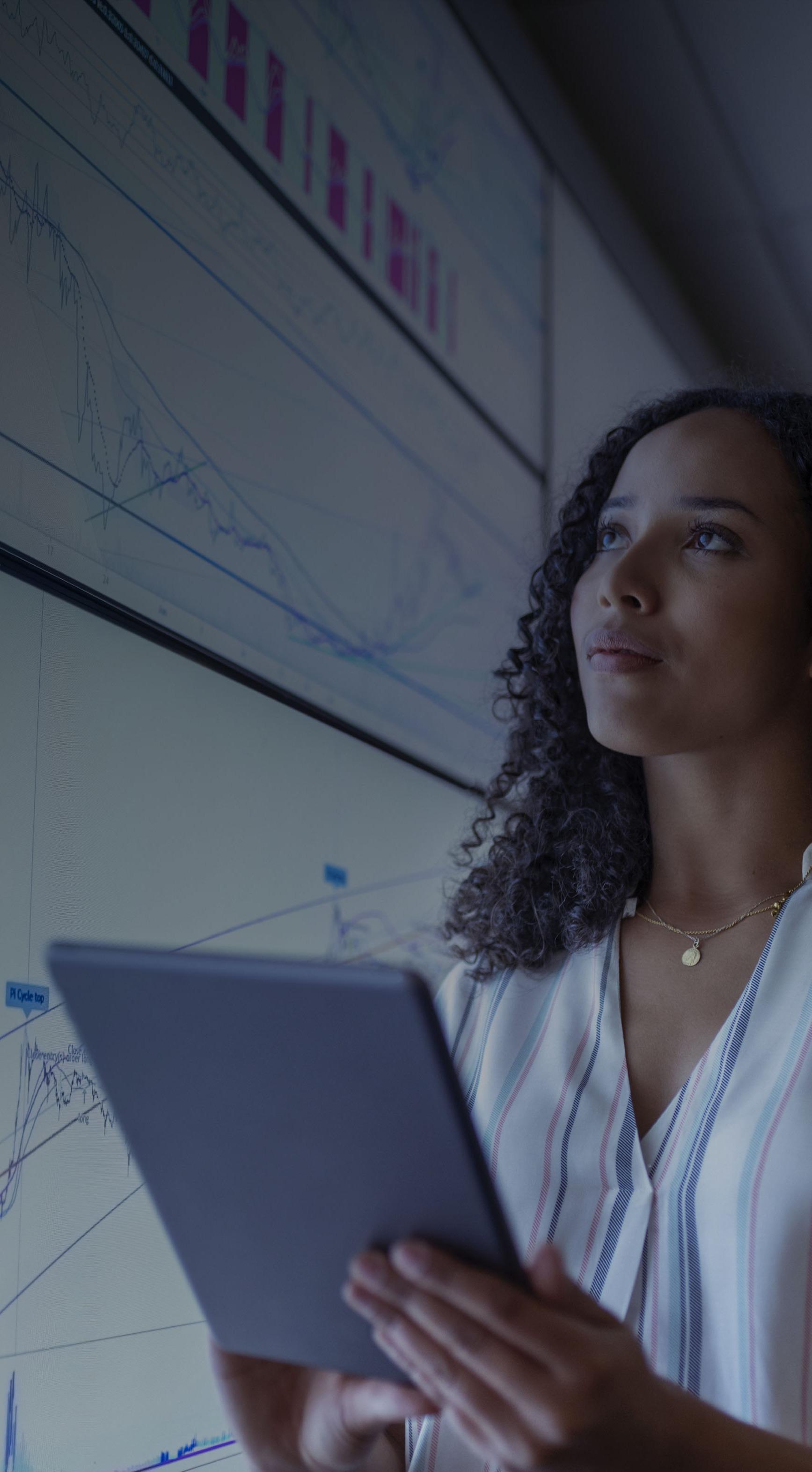 A woman stands holding a tablet, analyzing graphs displayed on a wall behind her.