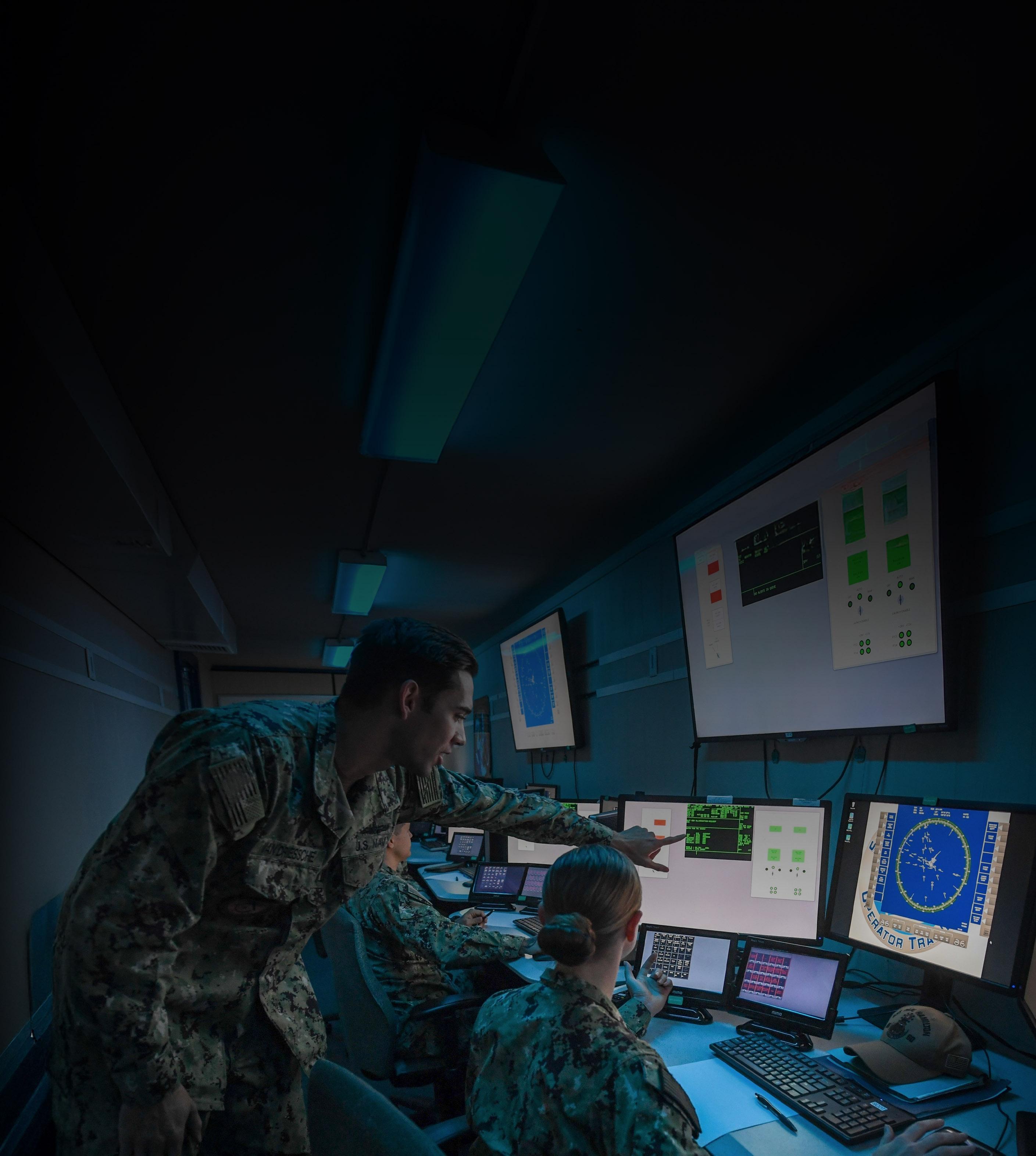 Military personnel focused on computers in a collaborative workspace, engaged in tasks and discussions.