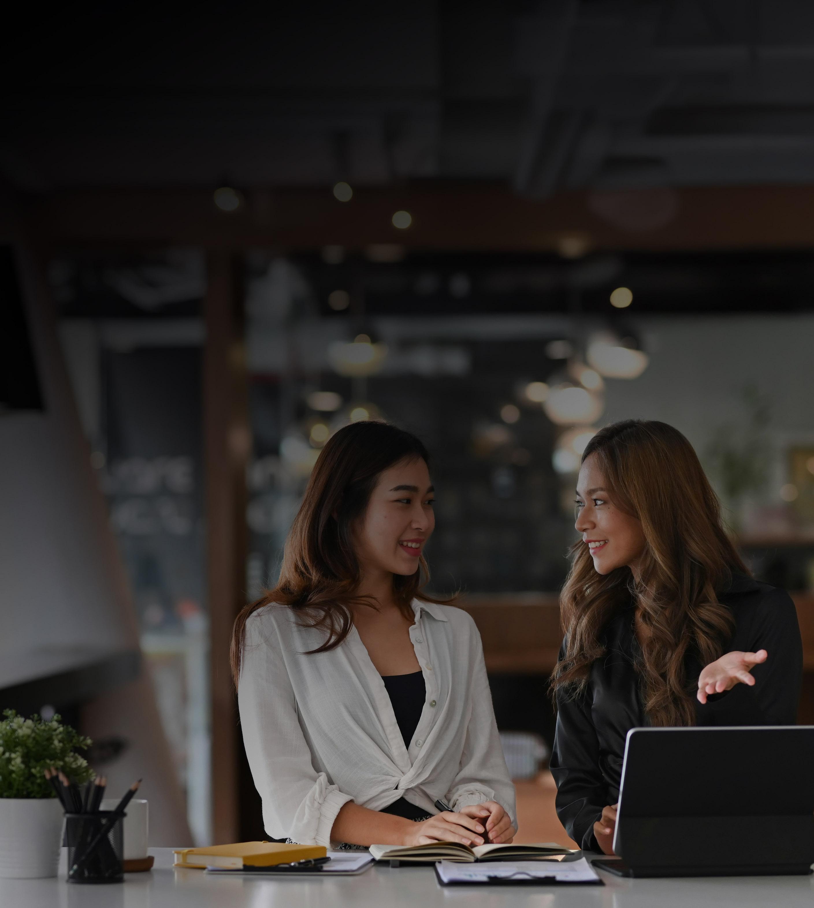 Two women seated at a table, collaborating on a laptop, engaged in discussion and sharing ideas.