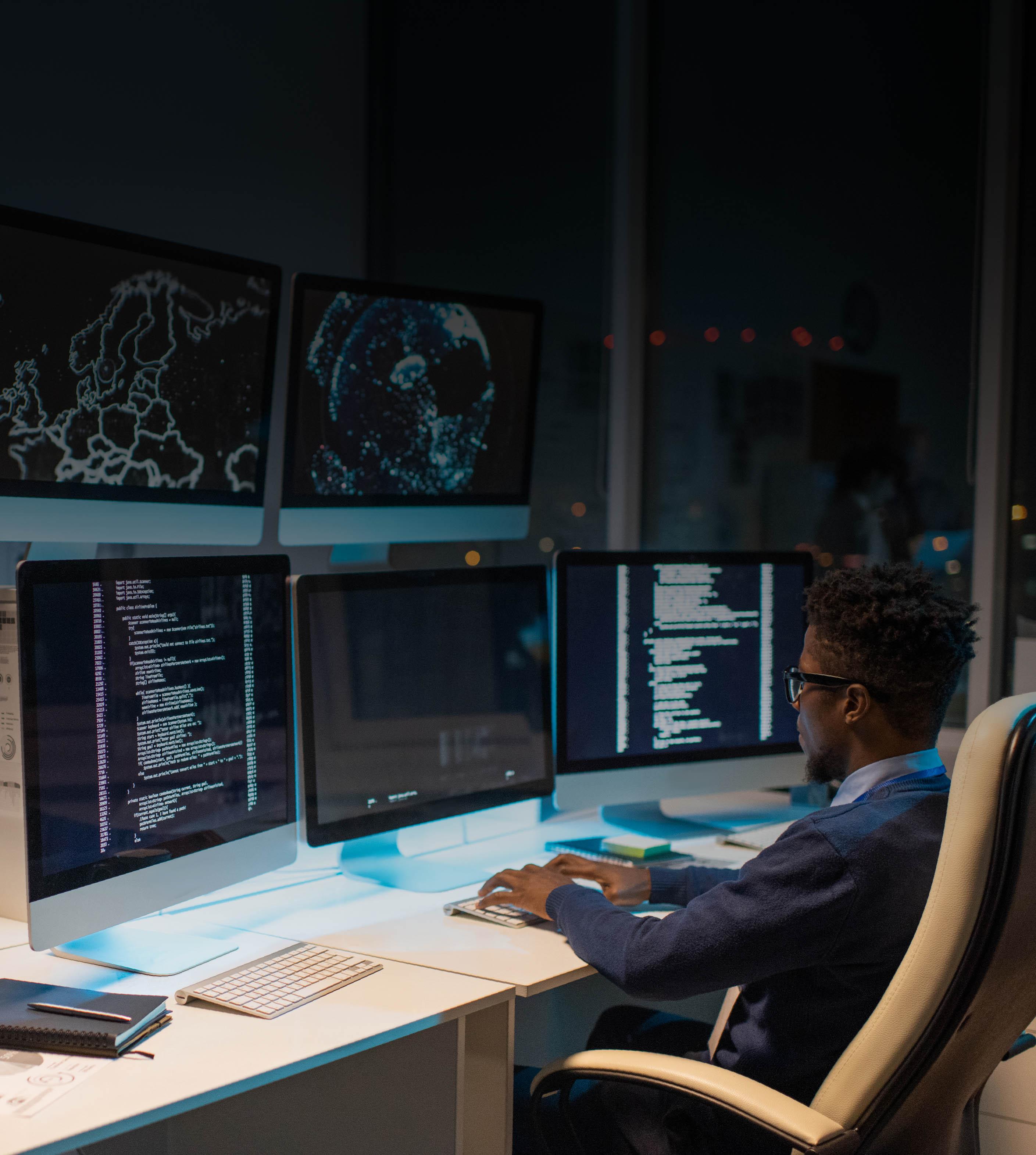A man seated at a desk, focused on three computer screens displaying various tasks and information.