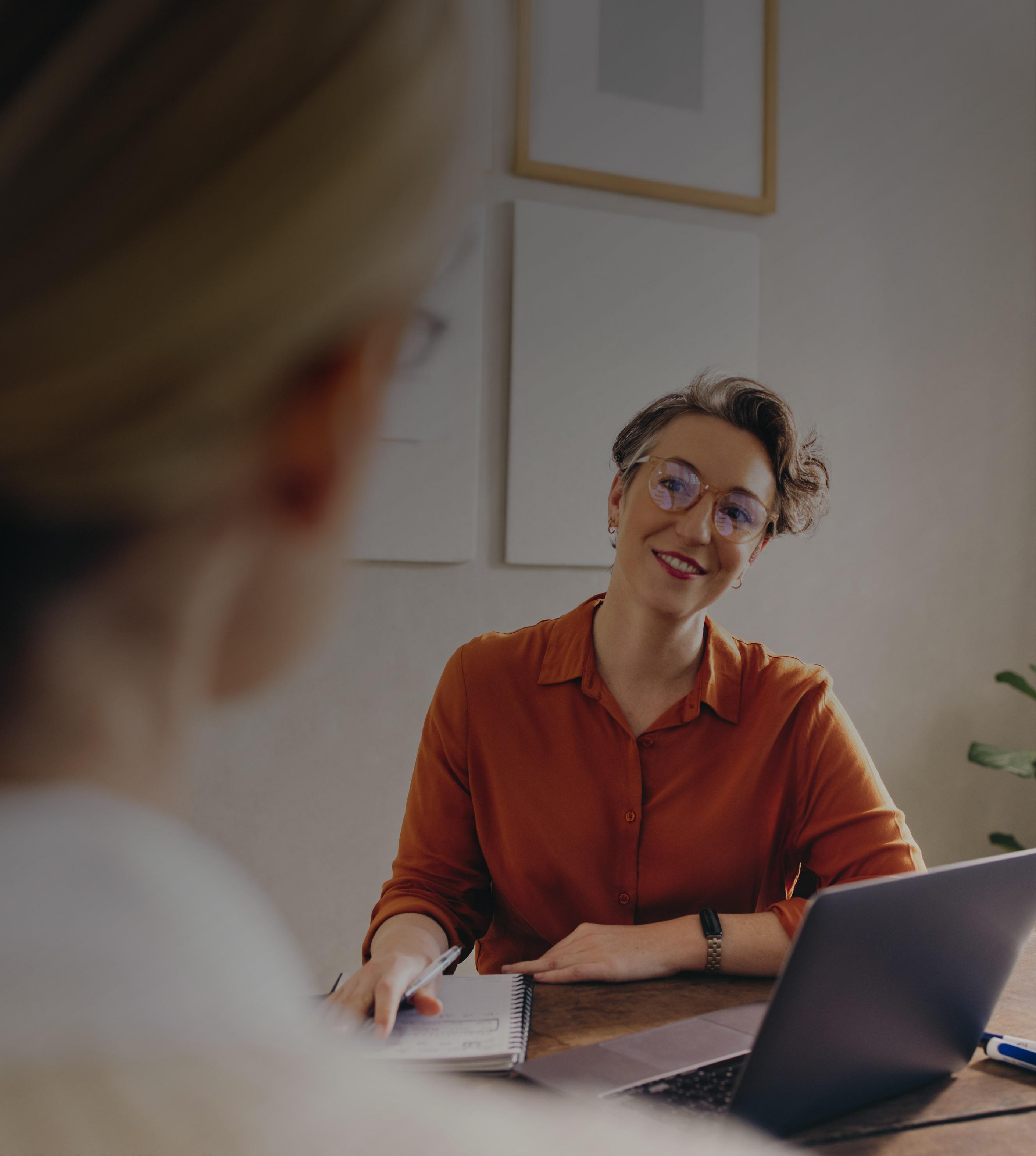 A woman seated at a desk, focused on her laptop, with a notepad and pen beside her.