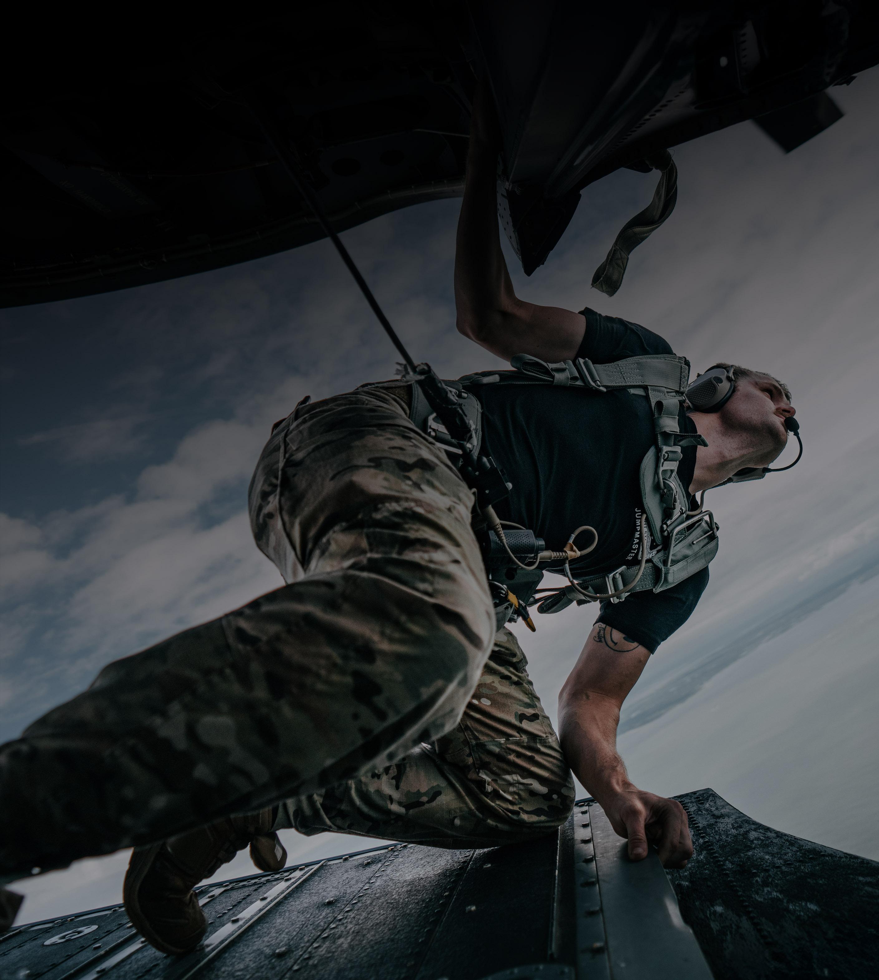 A man in military gear climbs out of a plane, preparing for a mission in a dynamic outdoor setting.