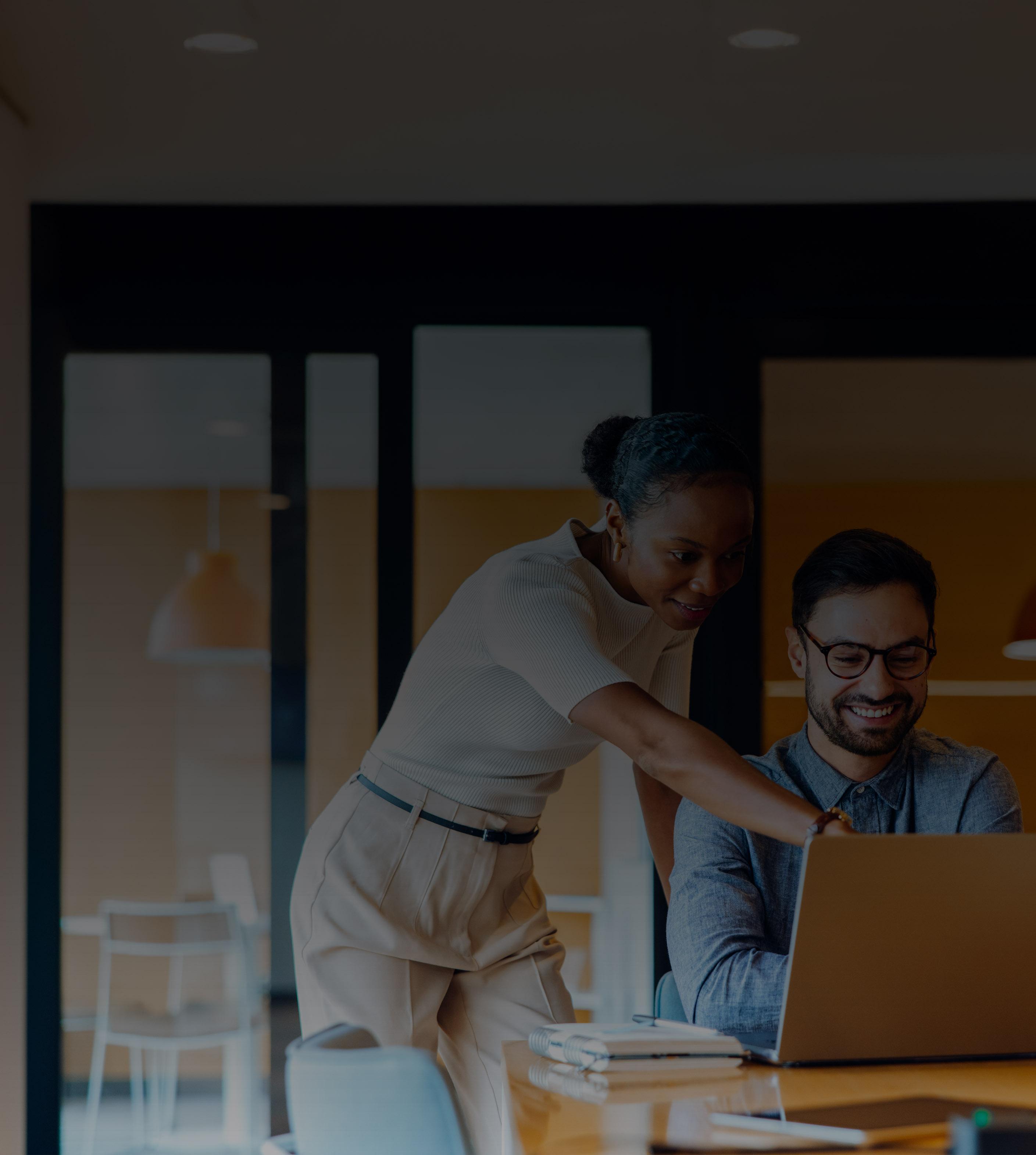A man and woman collaborate on a laptop in a modern office setting, focused on their work.