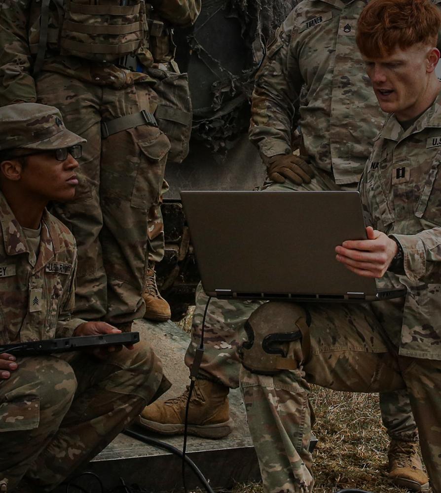 A group of soldiers gathered around a laptop, focused on the screen, engaged in a discussion or analysis.