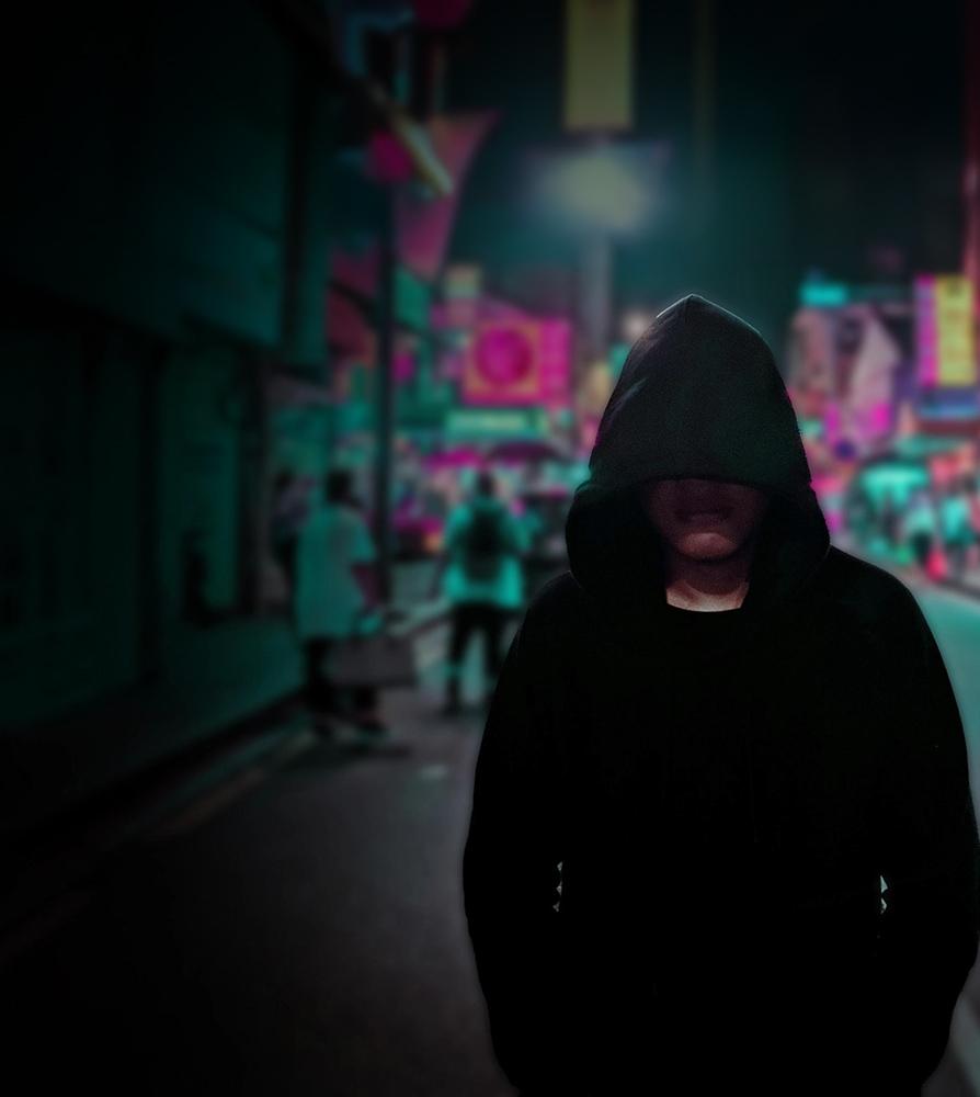 A person in a hoodie walking down a neon lighted street.