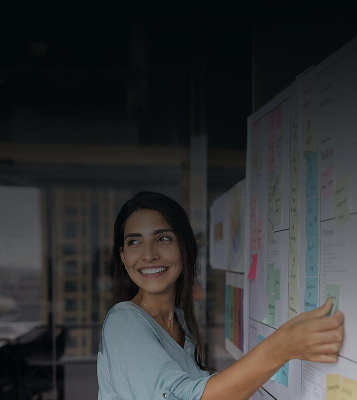 A woman points to a whiteboard covered in colorful sticky notes during a brainstorming session.