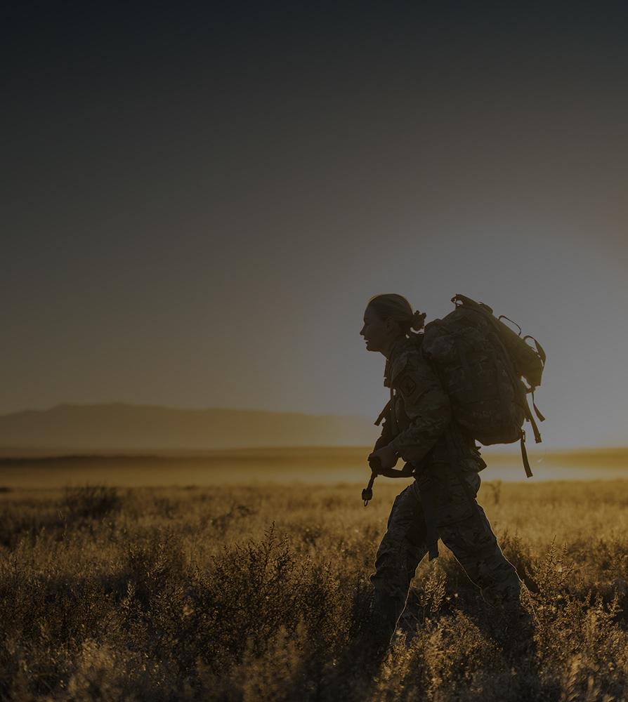 A woman man with a backpack walks through a field during sunset, with warm colors illuminating the landscape around him.
