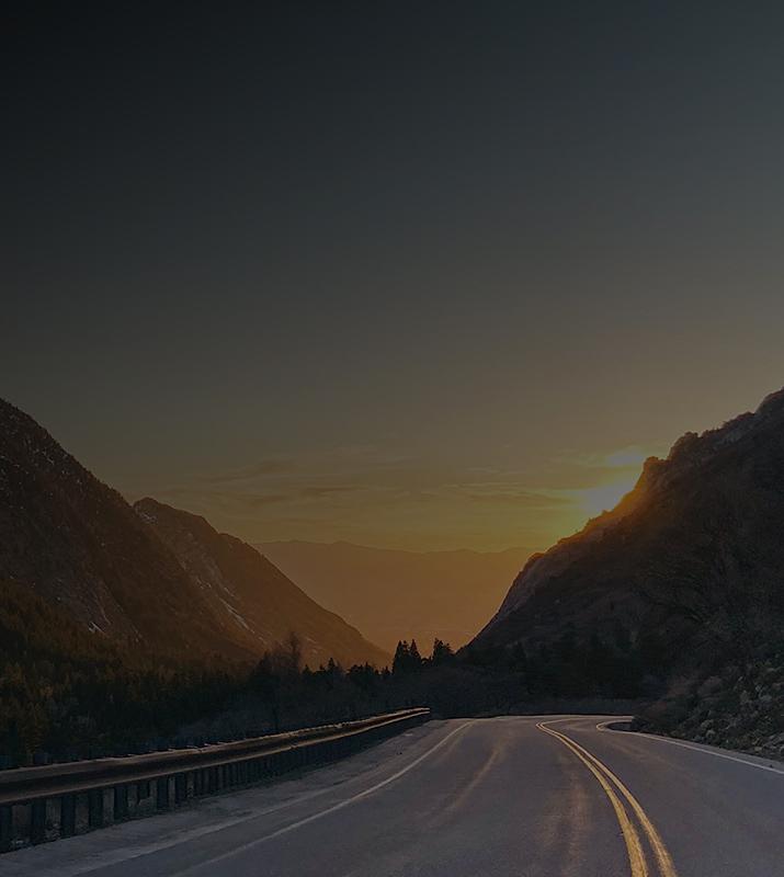 A scenic highway stretches into the distance, framed by majestic mountains under a vibrant sunset sky.