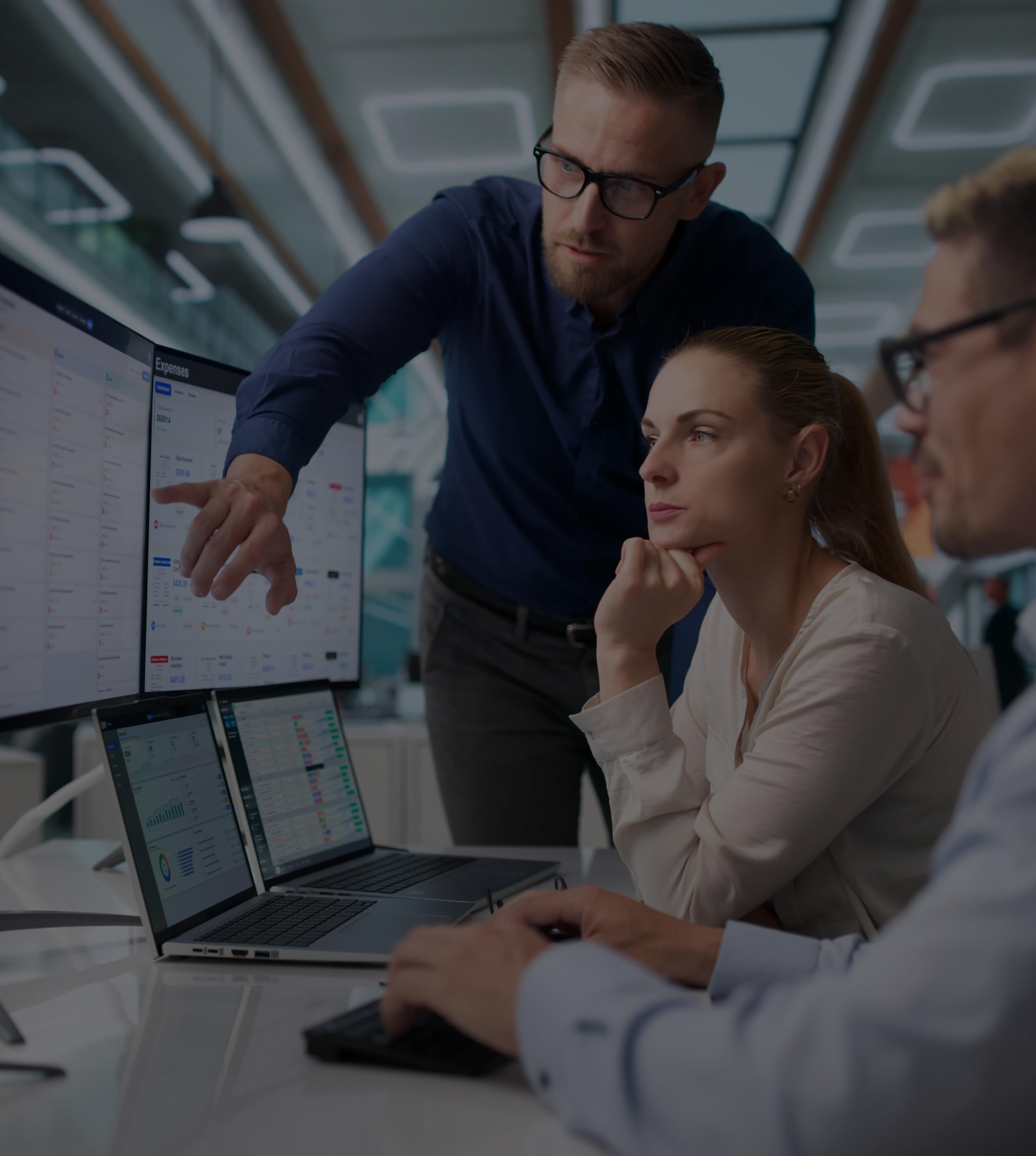 Three professionals collaborate at computer monitors in a modern office, focused on reviewing information in a serious, analytical mood.