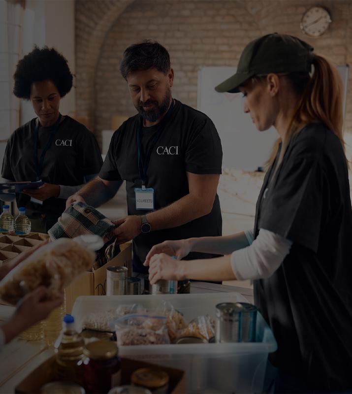 Volunteers prepare and serve food at a community table, working together in a warm kitchen with trays, bread, and drinks.