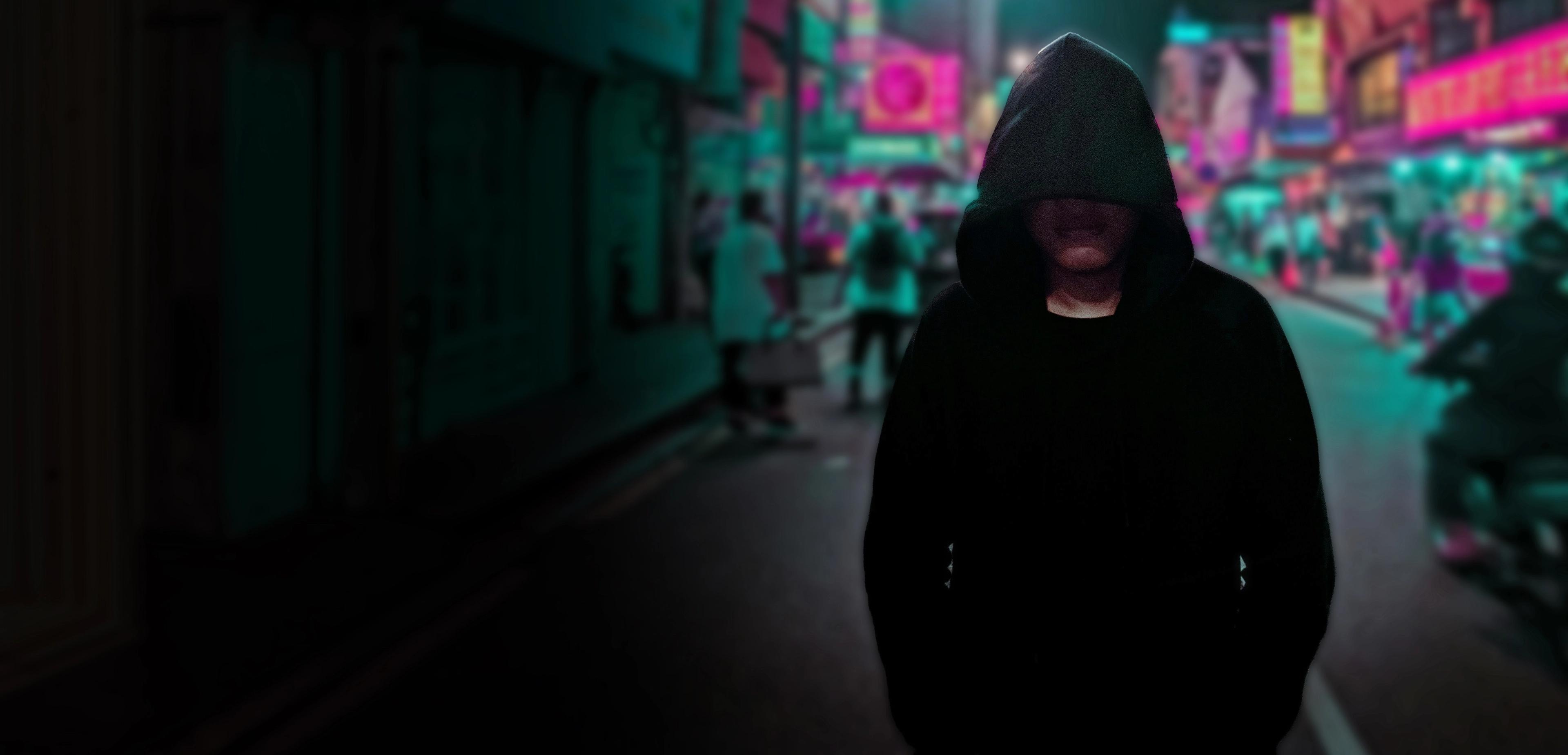 A person in a hoodie walking down a neon lighted street.