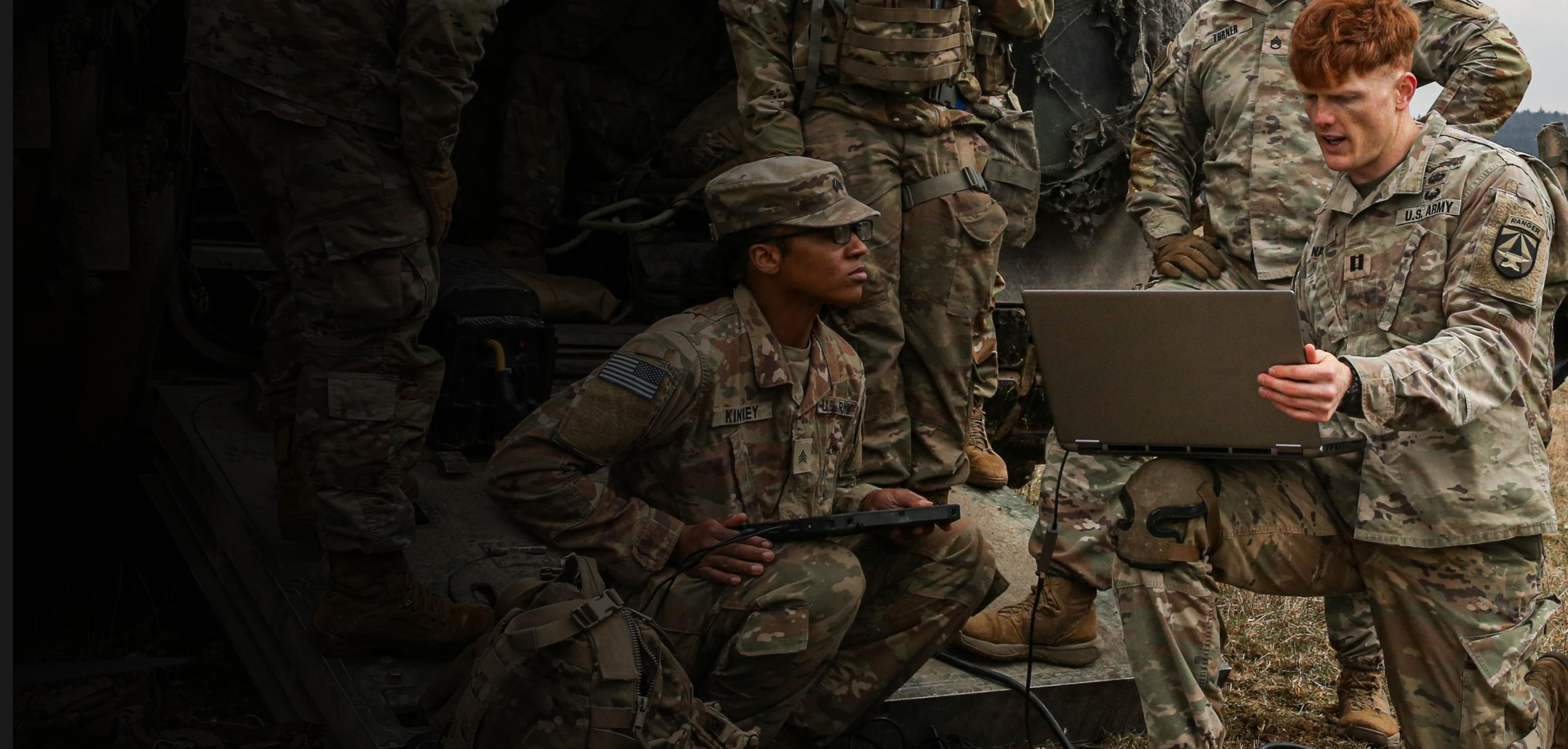 A group of soldiers gathered around a laptop, focused on the screen, engaged in a discussion or analysis.