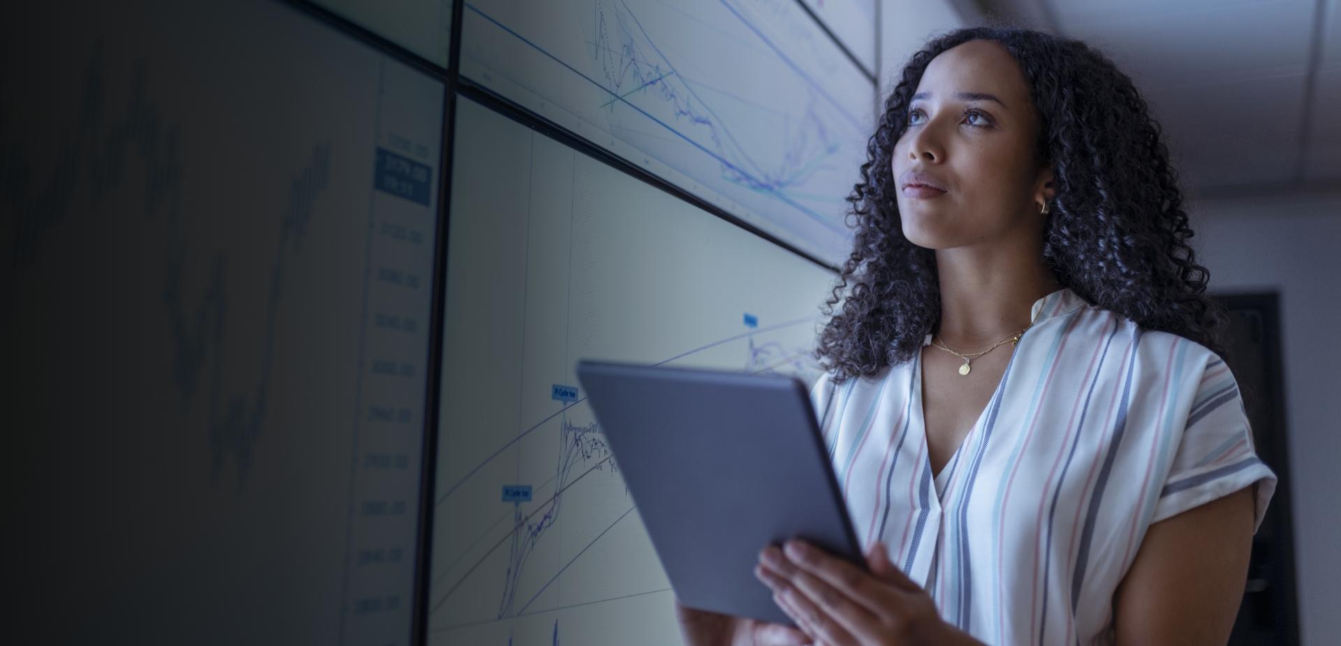 A woman stands holding a tablet, analyzing graphs displayed on a wall behind her.