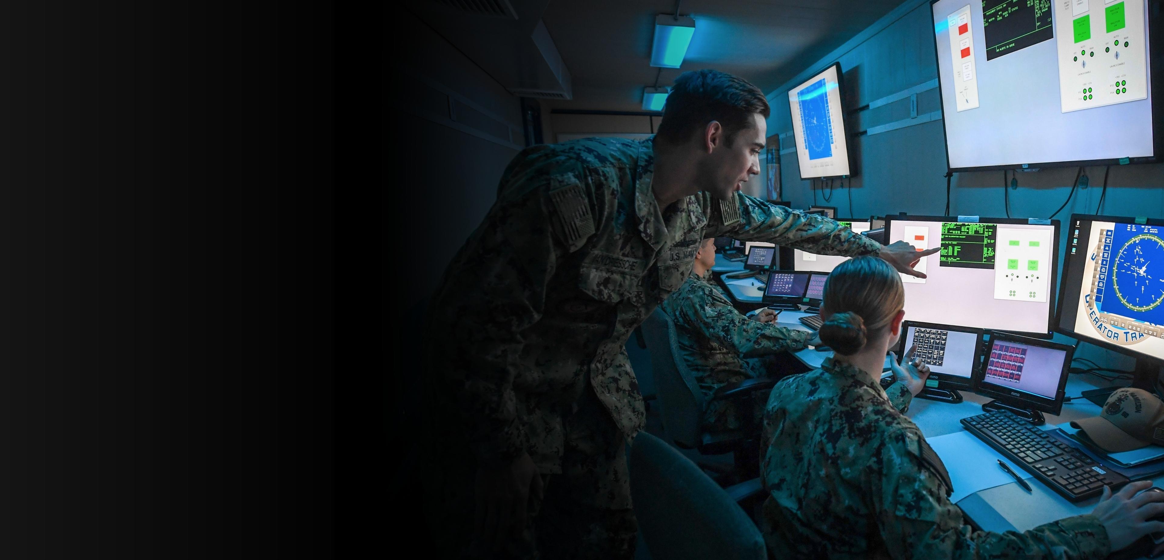 Military personnel focused on computers in a collaborative workspace, engaged in tasks and discussions.