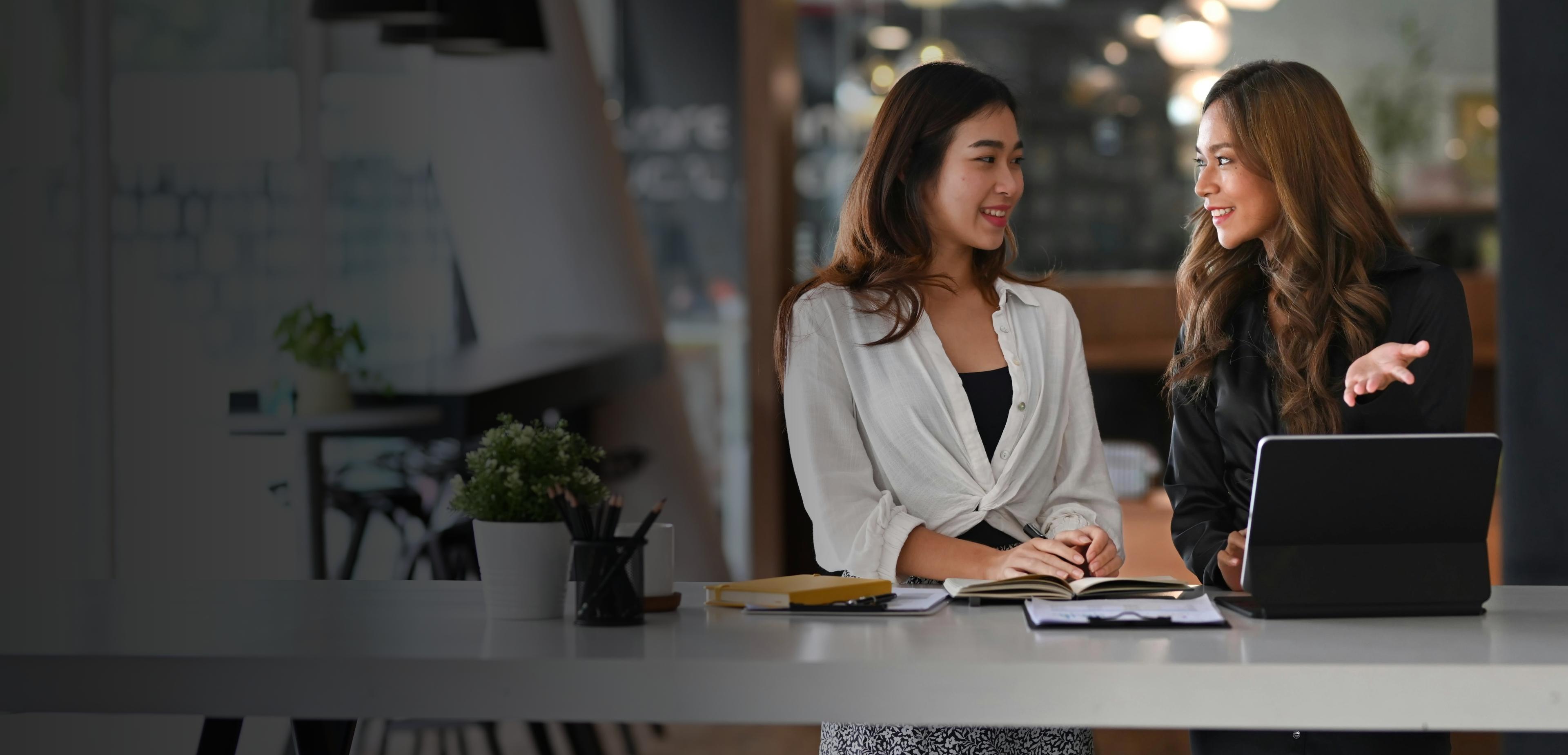 Two women seated at a table, collaborating on a laptop, engaged in discussion and sharing ideas.